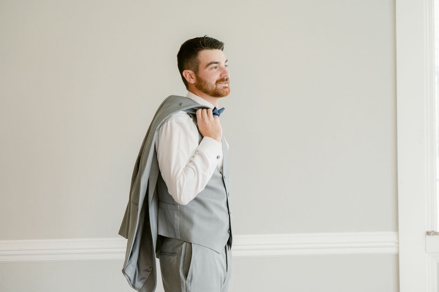 Central MN Wedding Photos | Groom in gray vest and navy bow tie holds jacket over shoulder in elegant wedding portrait.