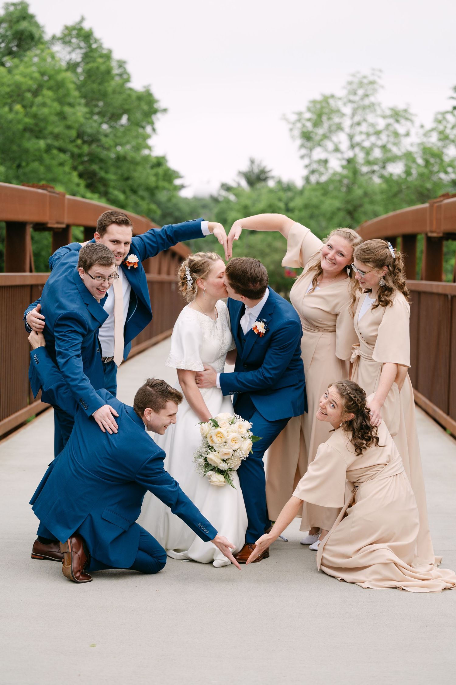 Wedding party having fun posing on wooden bridge with bride and bridesmaids in blush dresses and groomsmen in navy suits.