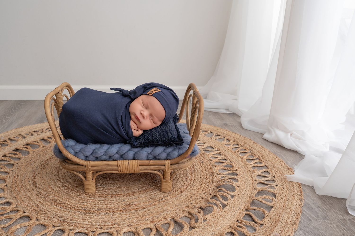 Minnesota Best newborn photographer - Sleeping newborn rests peacefully on woven rattan lounger atop circular jute rug near white curtains.