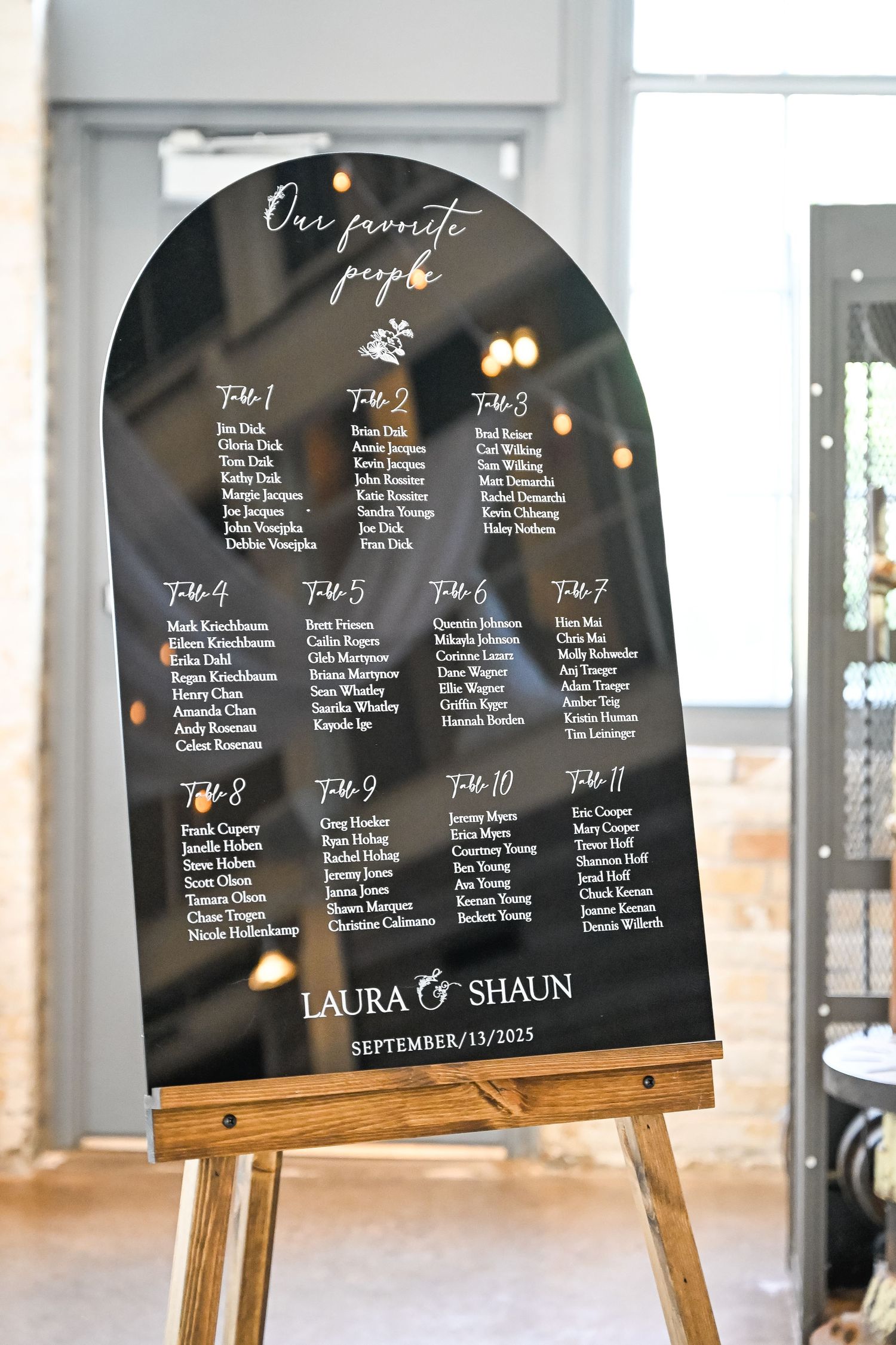 Black arch-shaped seating chart display on wooden easel shows wedding guest assignments.