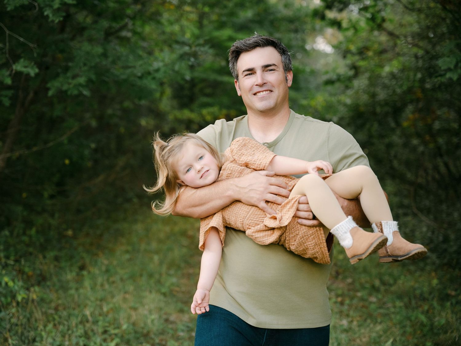 A smiling man holds a young girl and her teddy bear outdoors among lush green trees.. Minnesota milestone photographer
