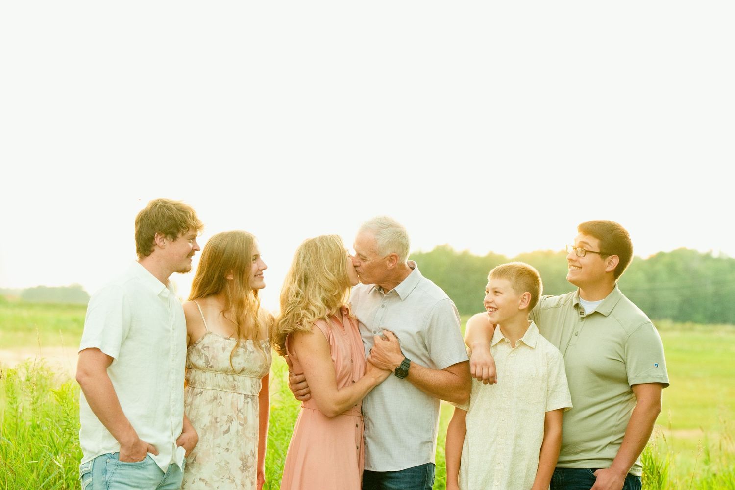 Family of six posing outdoors in a sunny green field, sharing a warm embrace and smiling together.