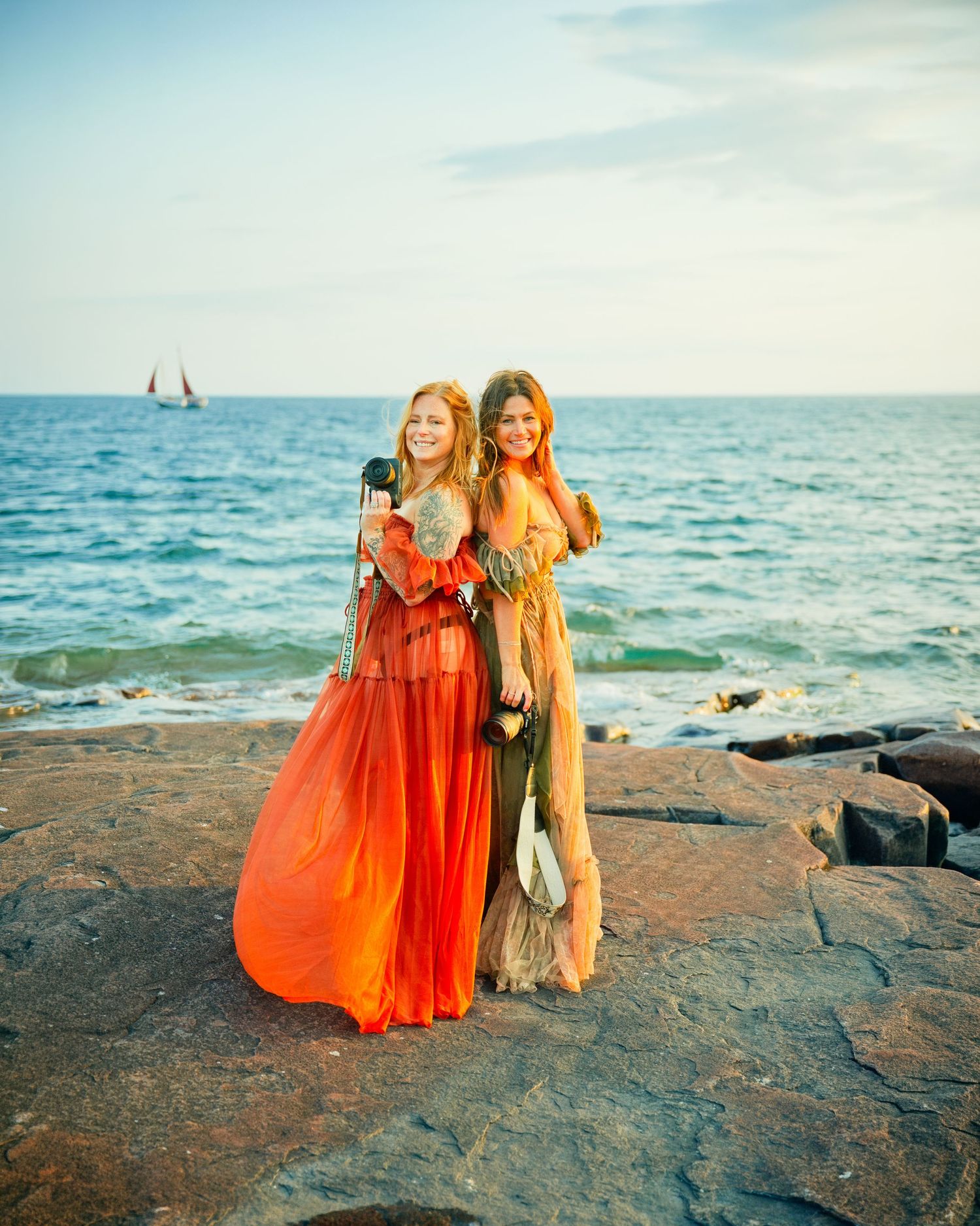Two women in flowing orange and gold gowns pose on rocky shoreline with sailboat and blue ocean backdrop.