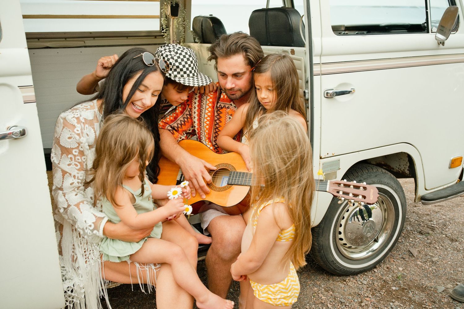 Bohemian family with children gathered around a man playing acoustic guitar by a vintage camper van outdoors.