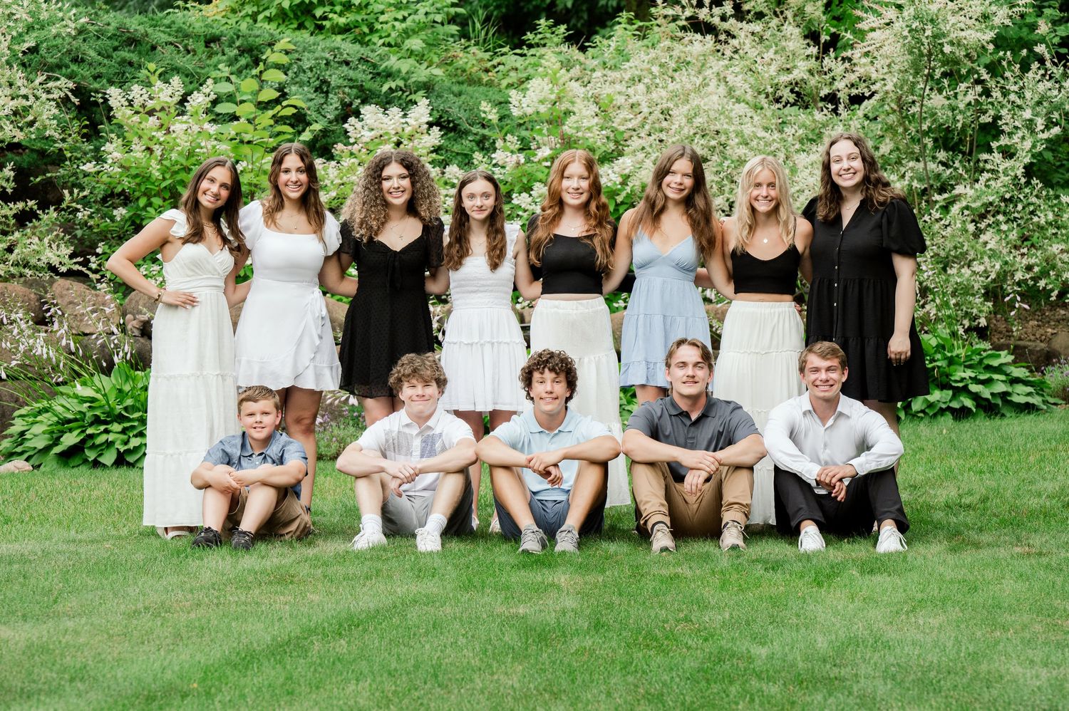 Mixed group in white dresses and casual attire posing together in outdoor garden setting.