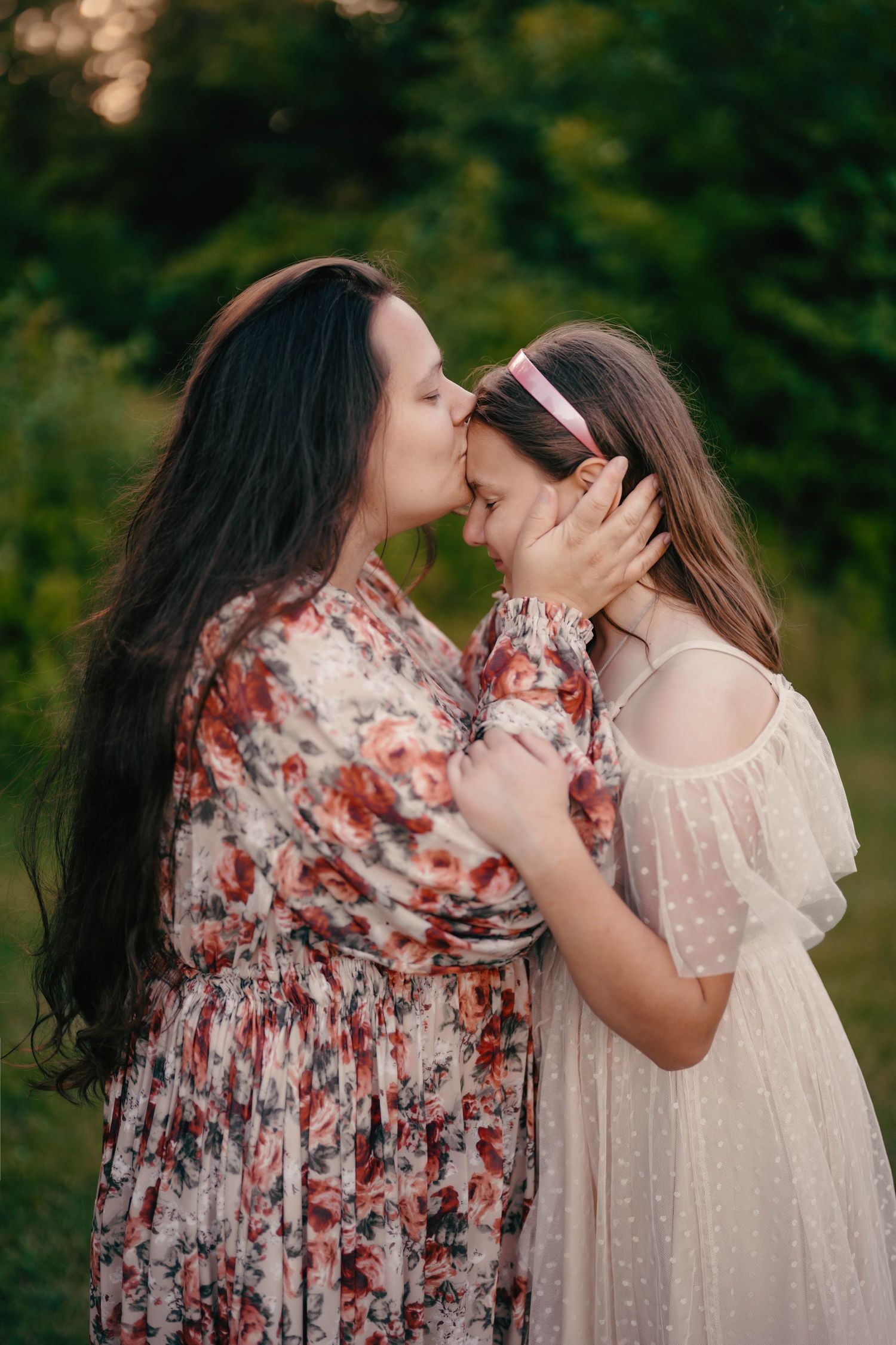 Two people in floral and light dresses embrace tenderly against a green natural backdrop.