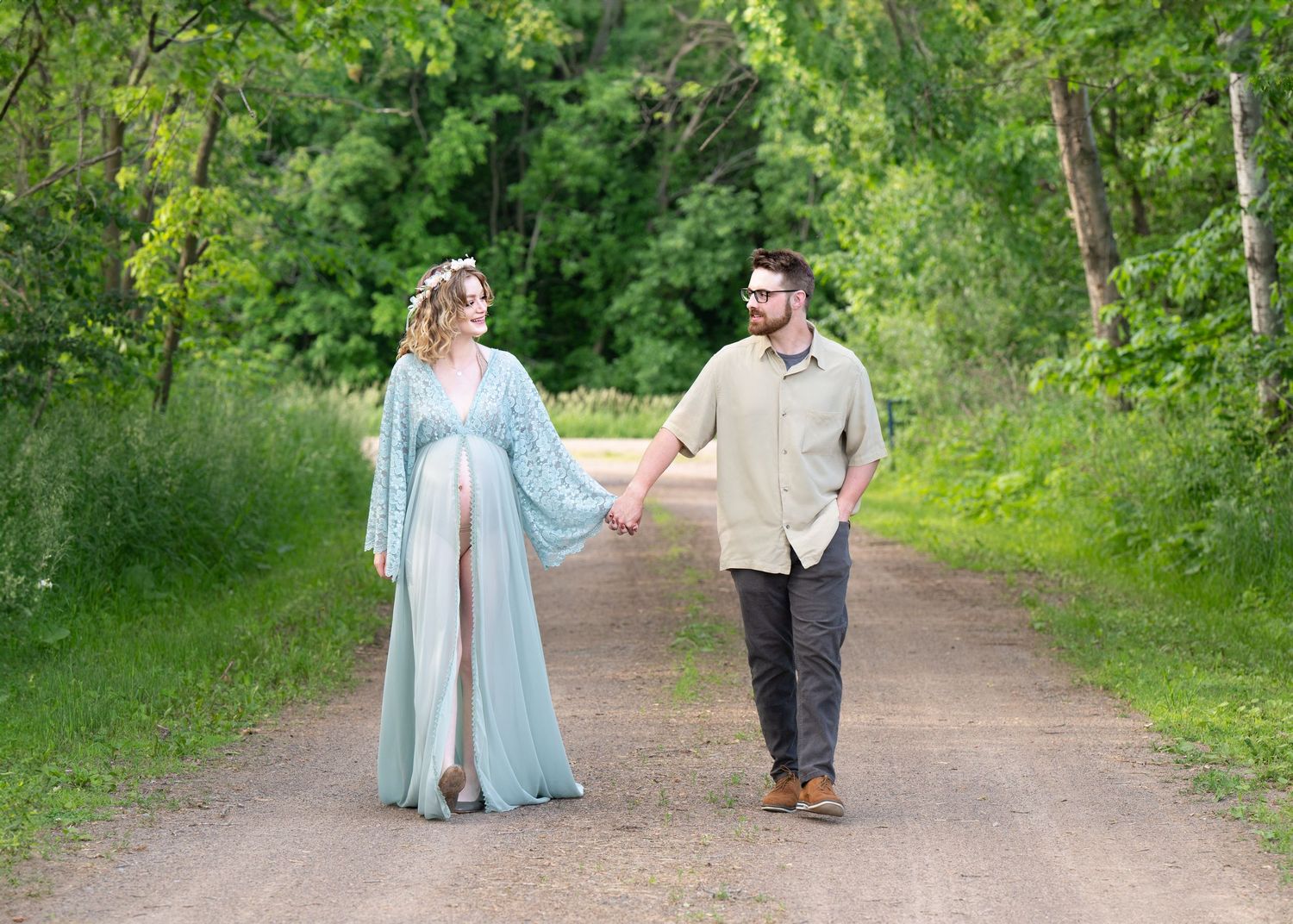 A couple walks hand in hand on a tree-lined dirt path during a summer maternity photoshoot. Princeton MN Milestone, Maternity and Newborn P