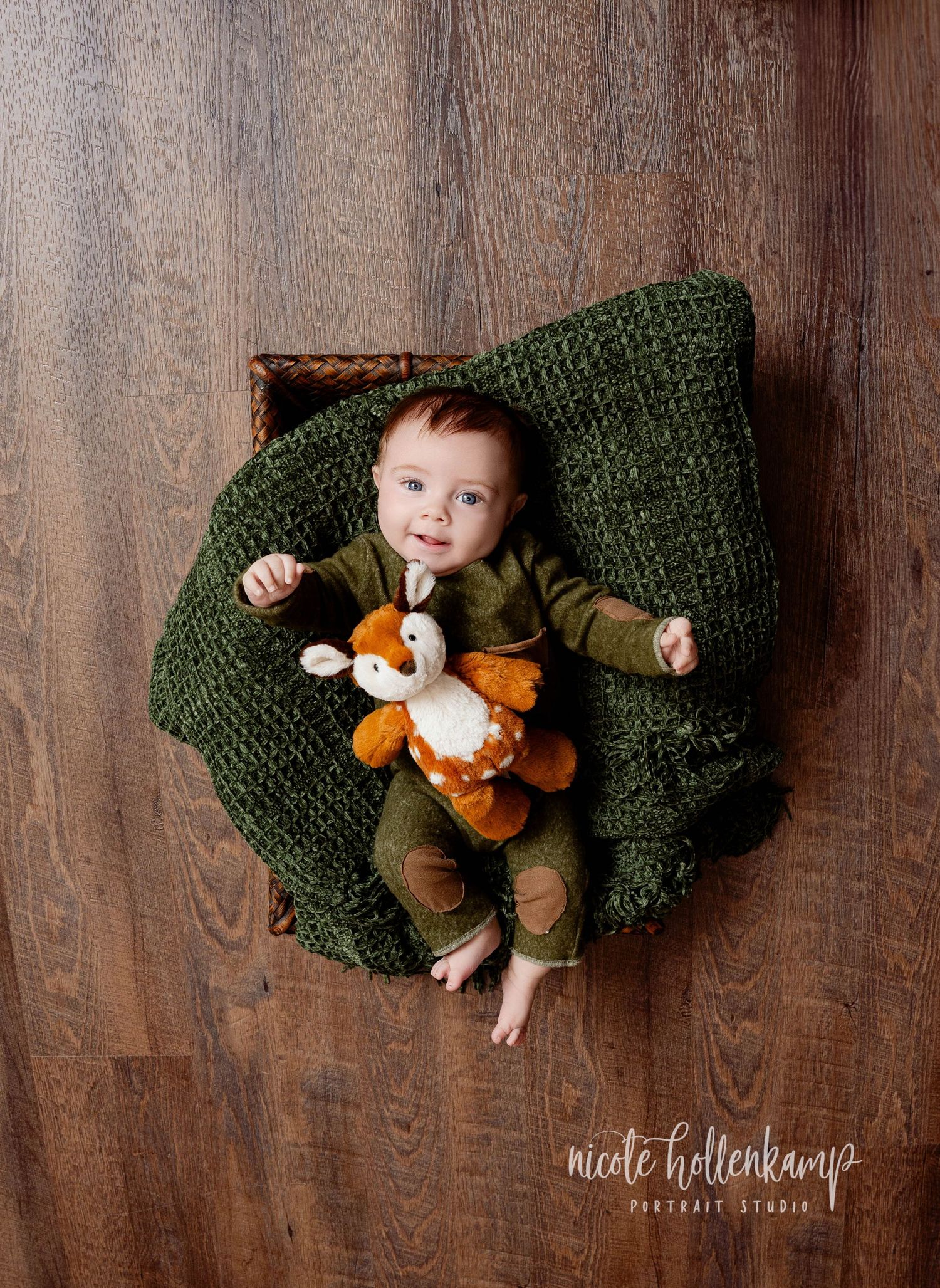 A baby in brown clothing lies on a chunky knit green blanket with an orange plush fox toy against a wooden background.