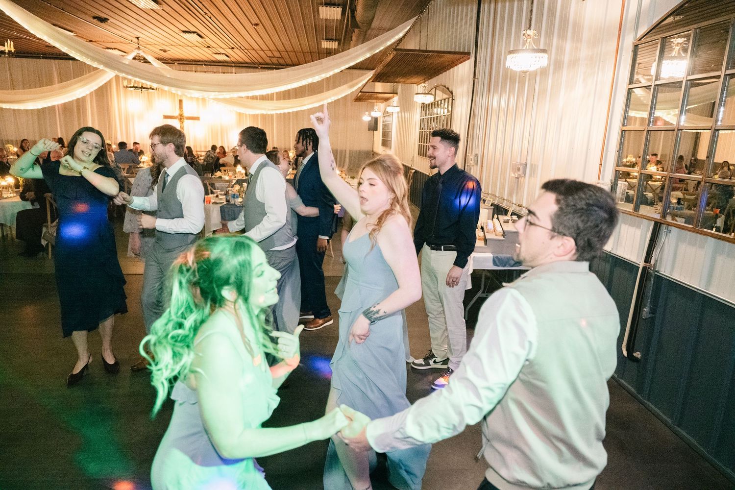 Guests dancing joyfully under colorful lights at a rustic wedding reception.