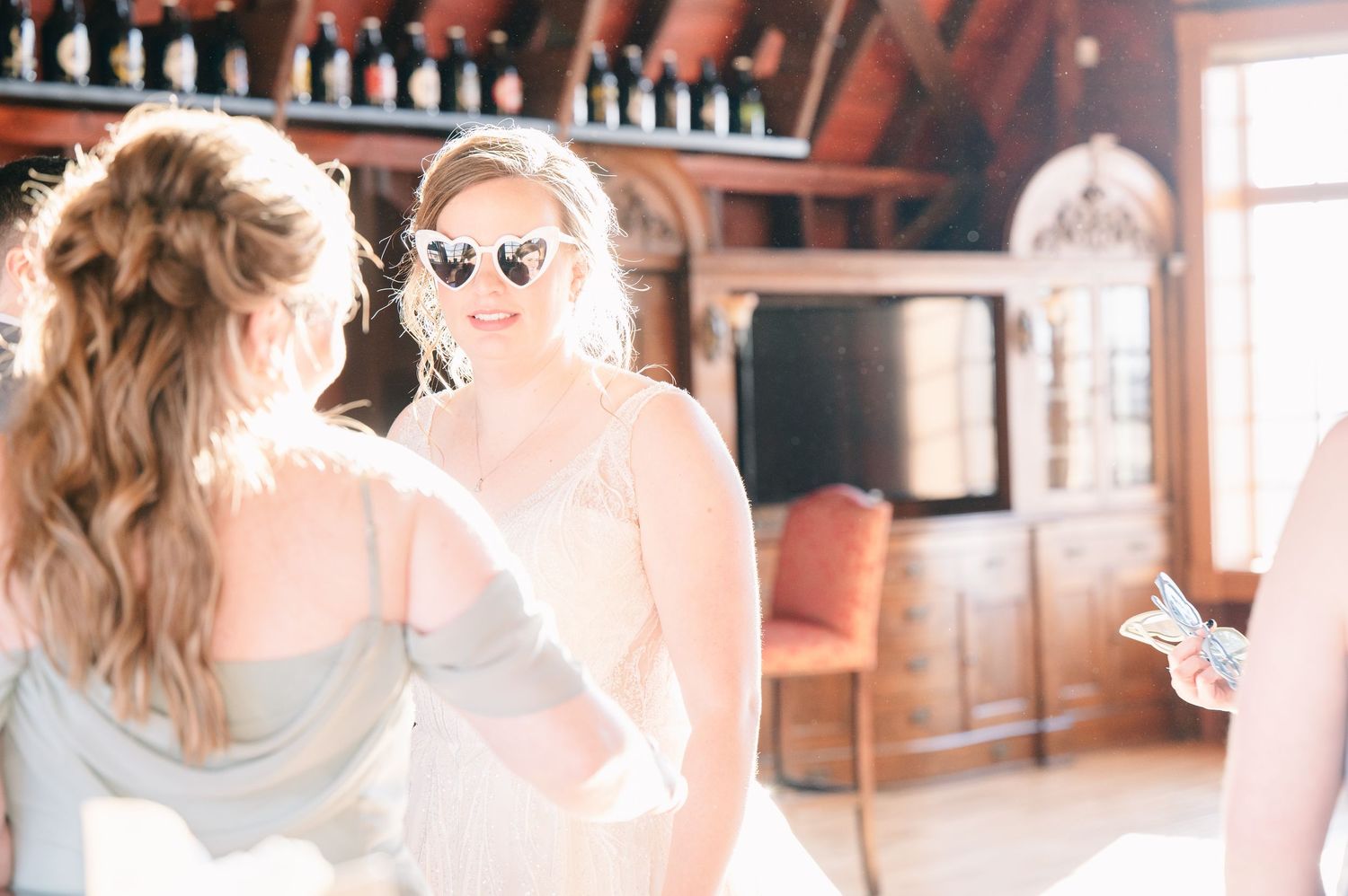 Bridesmaid in gray dress wearing heart-shaped sunglasses smiling in sunlit rustic venue interior.