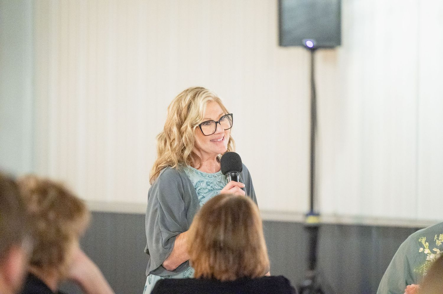Blonde woman with glasses gives a heartfelt speech at a wedding reception while holding a microphone.