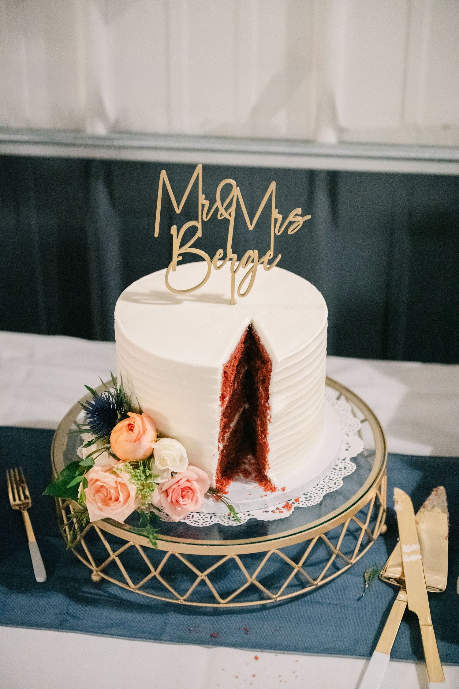 Two-tier white wedding cake with Mr and Mrs Berge gold topper, sliced to reveal red velvet interior.