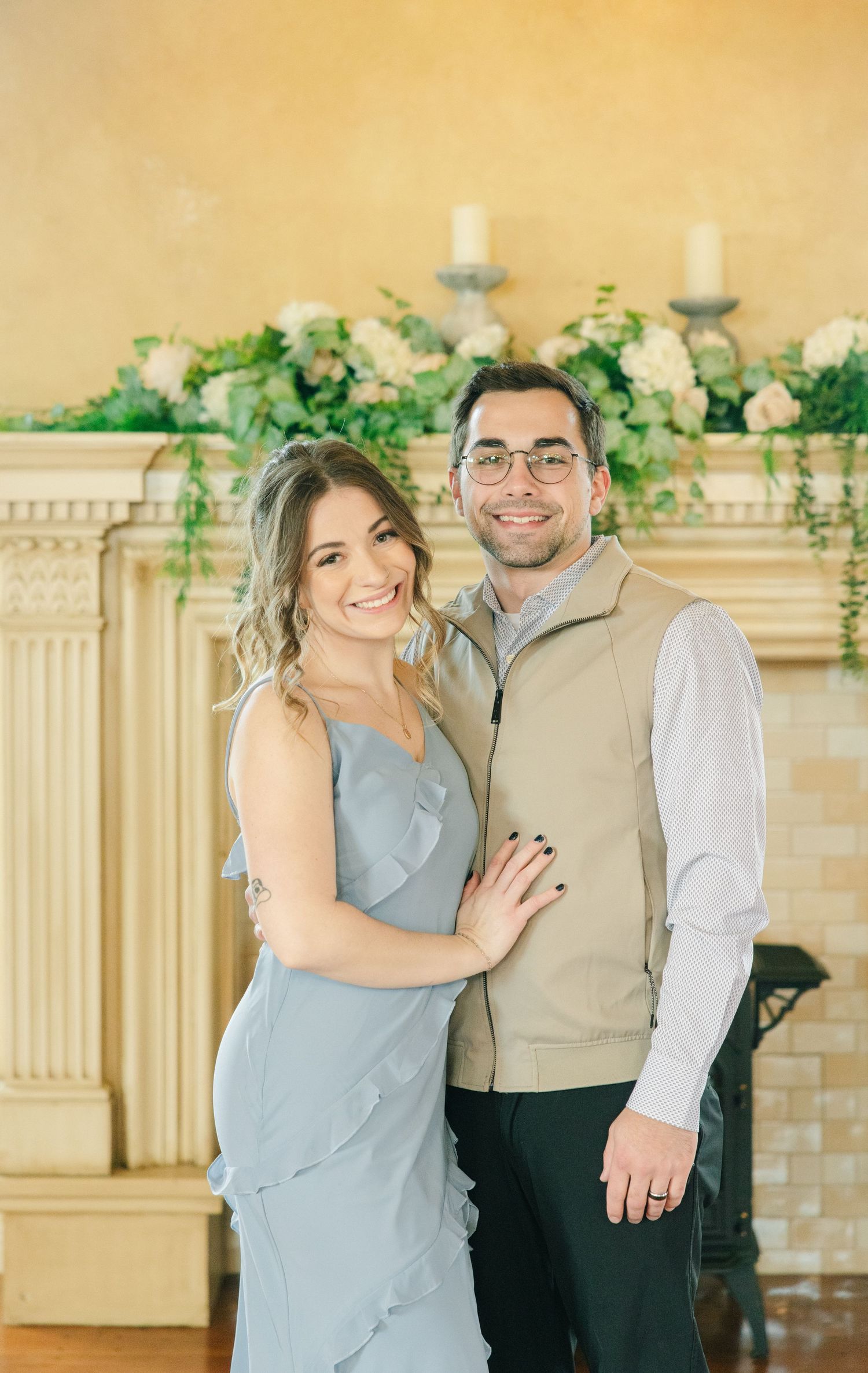 Smiling couple poses together in front of a floral fireplace mantle, woman in blue dress, man in tan vest.