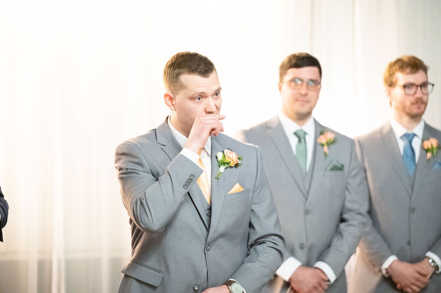 Emotional groom covers his mouth while waiting at the altar with groomsmen in gray suits during wedding ceremony.