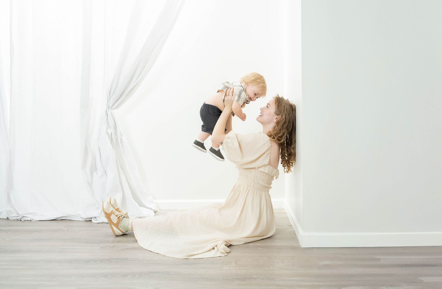 A parent in a flowing white dress lifts a baby joyfully in a bright minimalist studio setting.