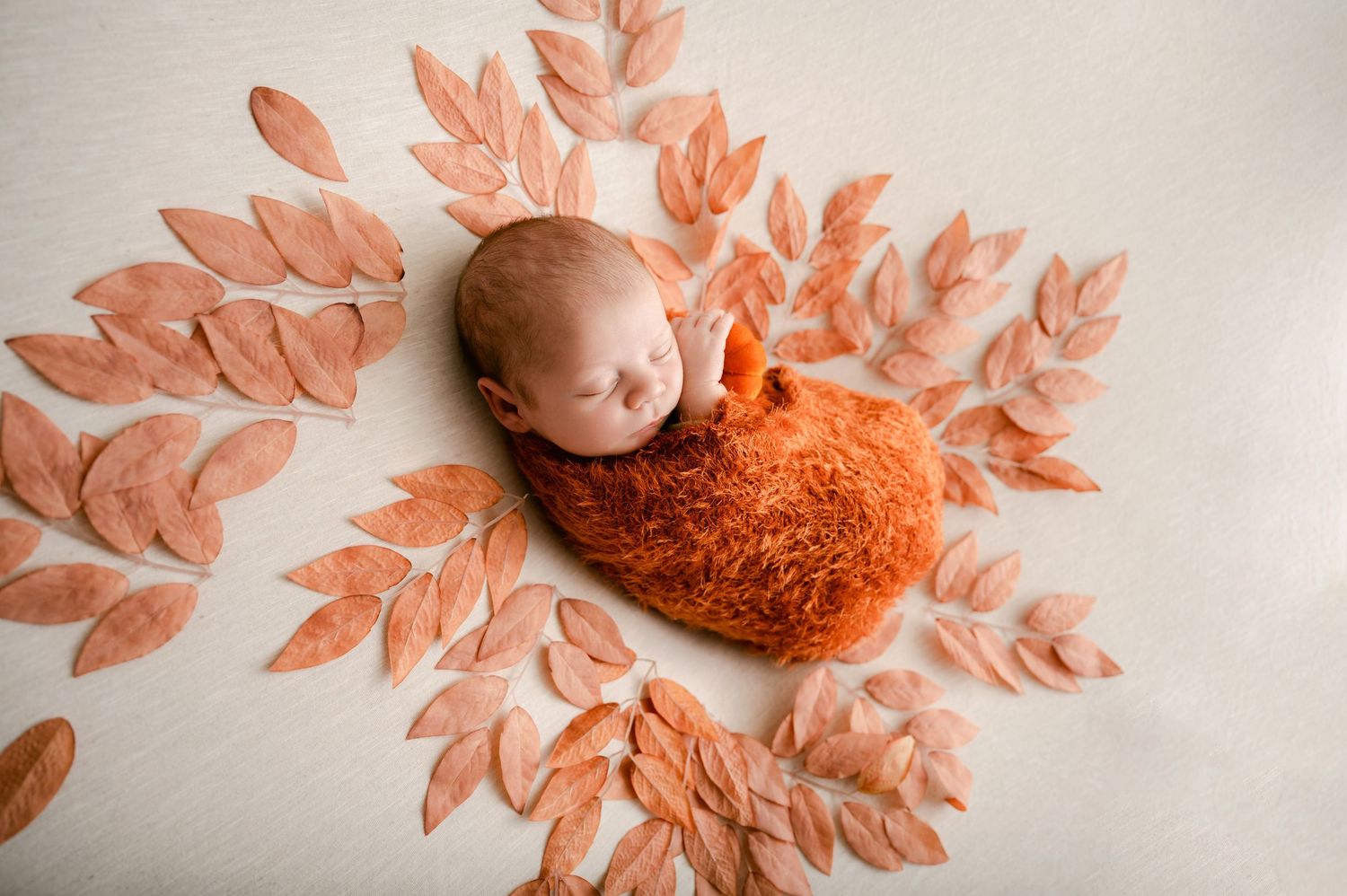 Newborn sleeping peacefully on orange blanket surrounded by peach-colored leaf decorations.