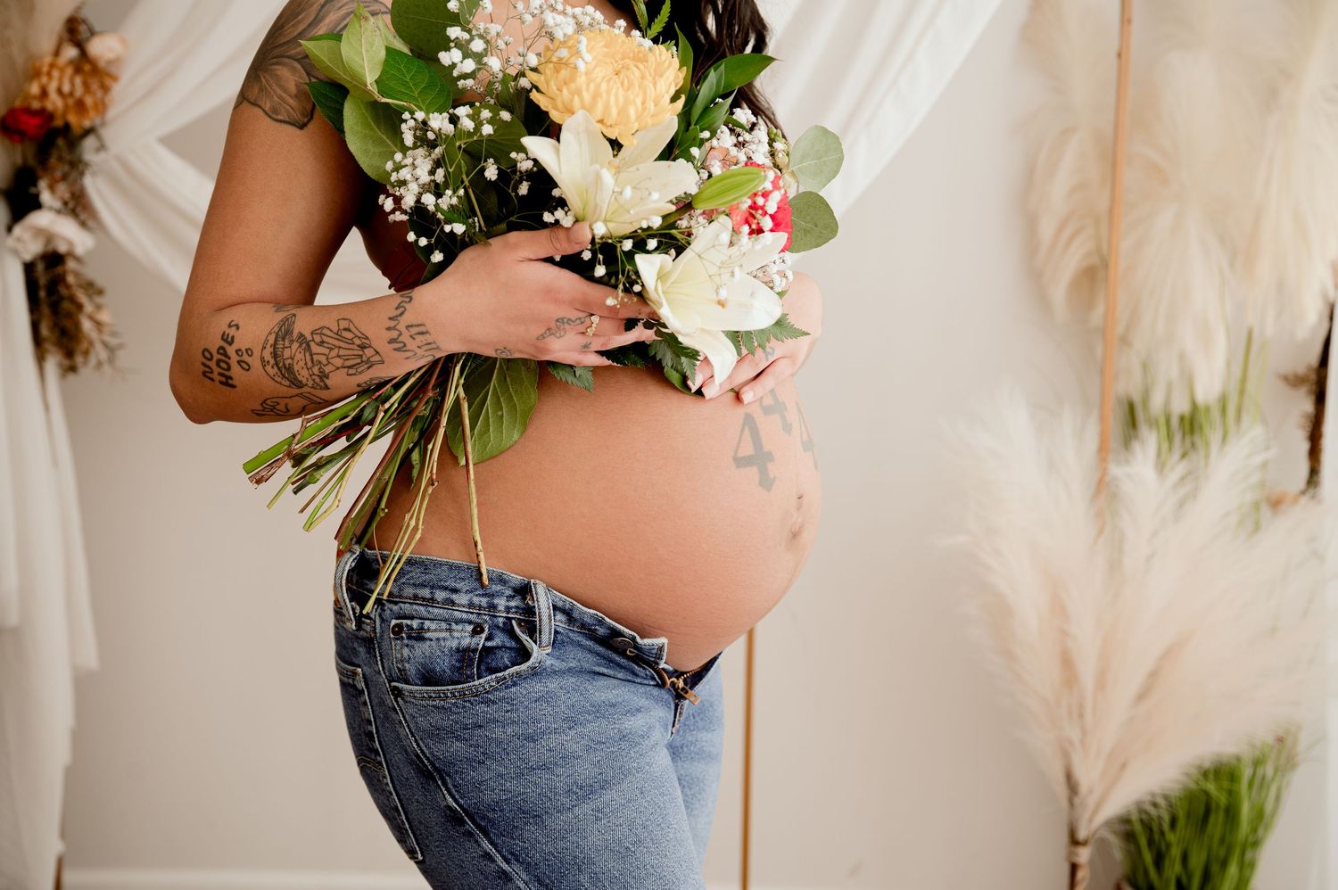 A pregnant belly in blue jeans holding a delicate bouquet of white roses and assorted flowers against a light backdrop.