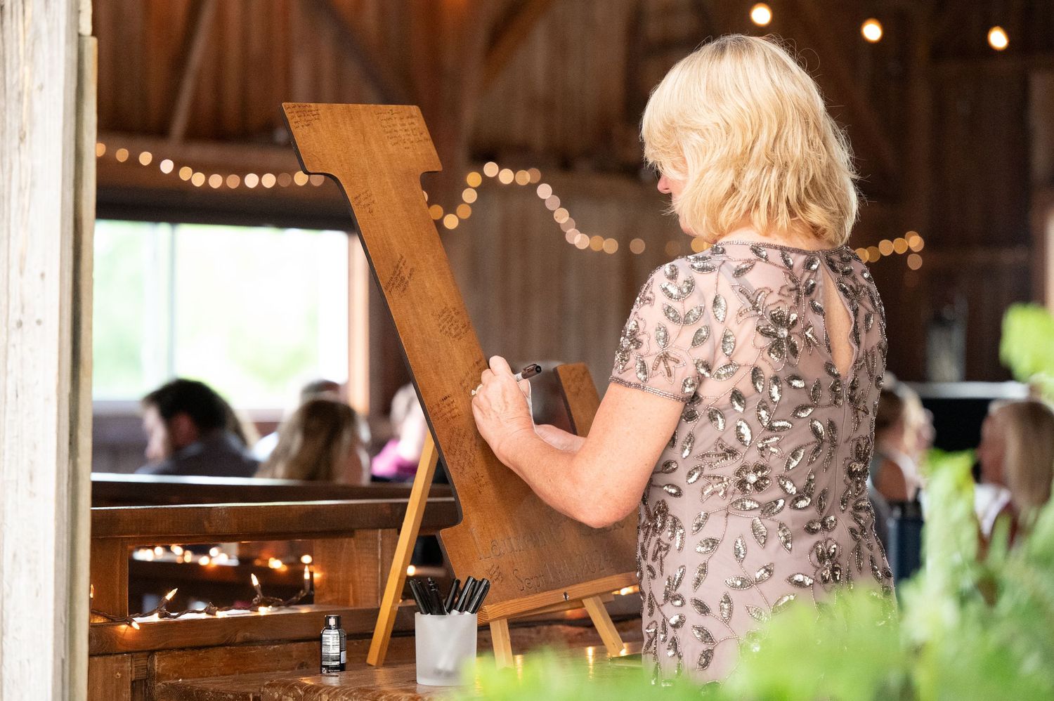 A person in a floral dress stands at a wooden letter display with string lights decorating a rustic barn venue.