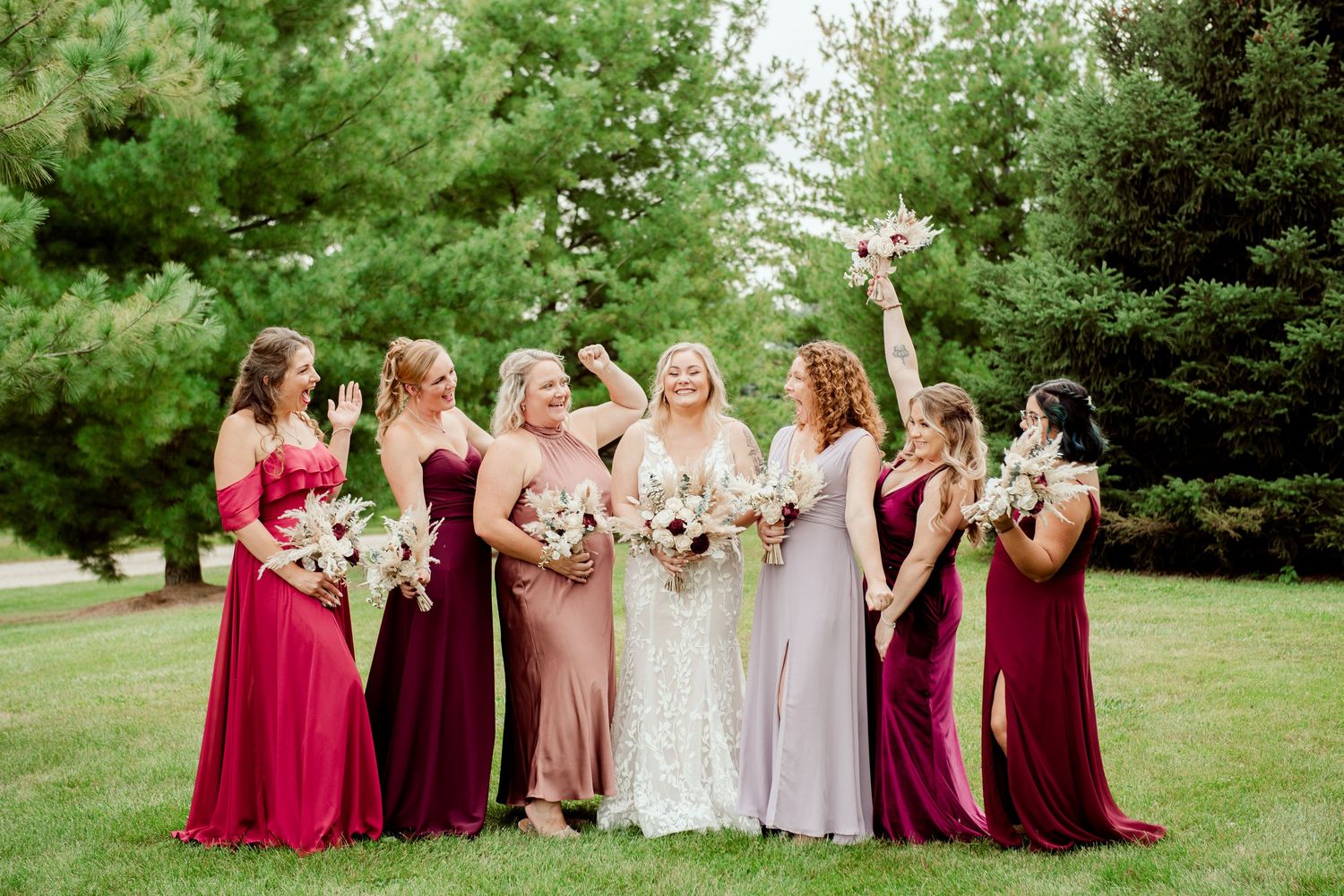 Bridesmaids in burgundy and blush dresses pose on a lush green lawn.