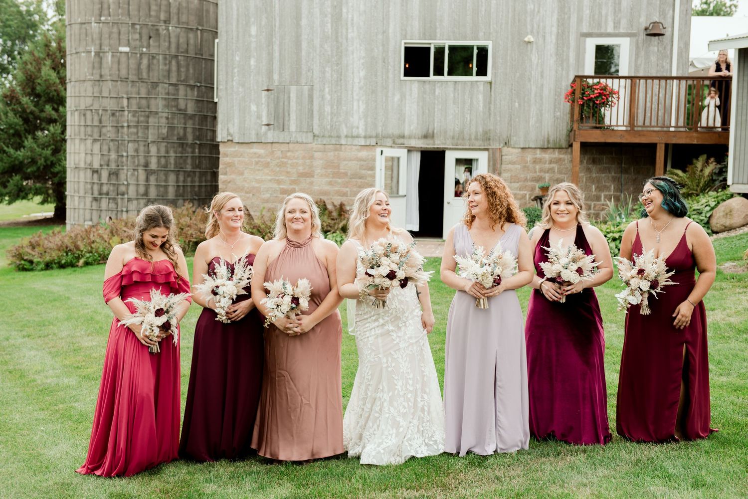 A bridal party wearing burgundy and blush dresses poses together in front of a rustic barn.