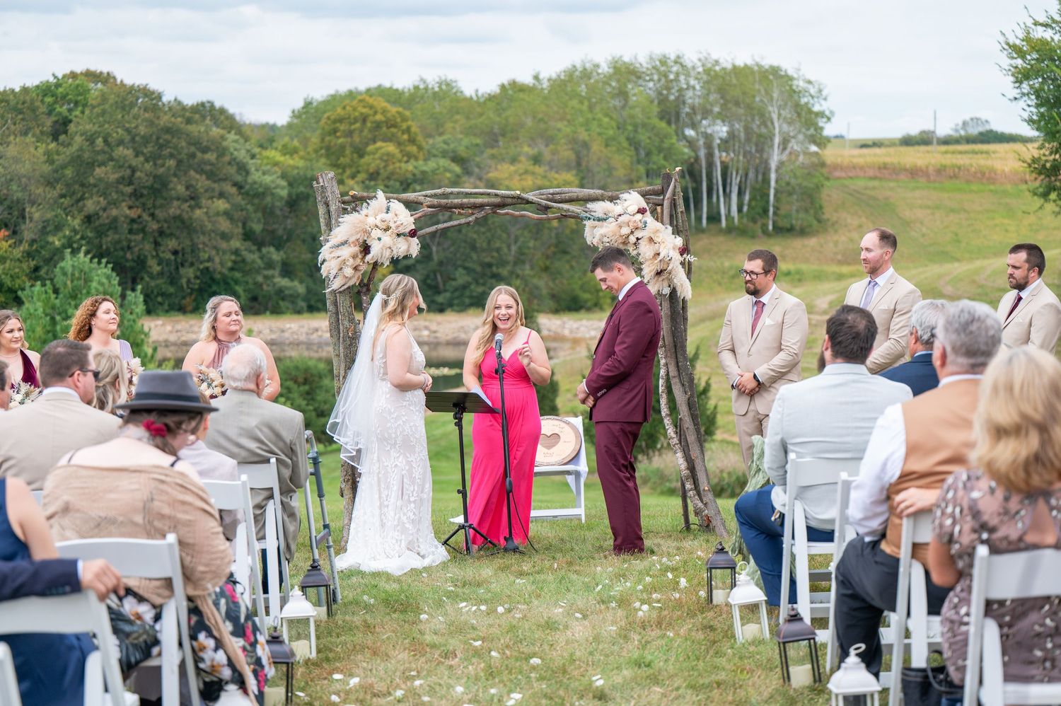 Wedding ceremony in progress with guests seated outdoors under floral arch on hillside.