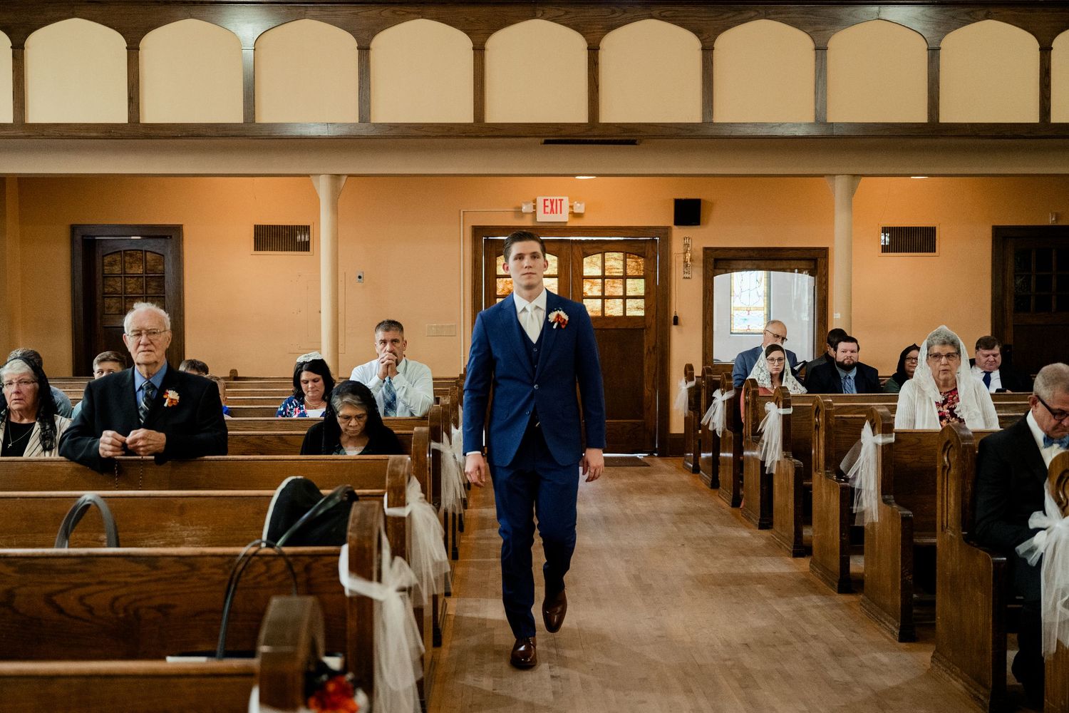 A person walks down the center aisle of a traditional church with wooden pews filled with wedding guests.