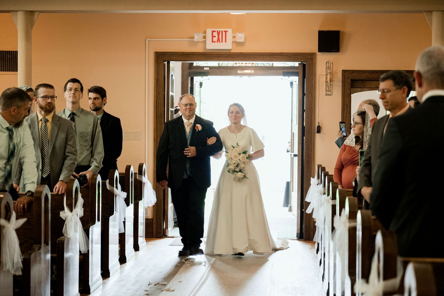 Traditional church wedding entrance with beautiful long white dress walking down wooden pew-lined aisle.