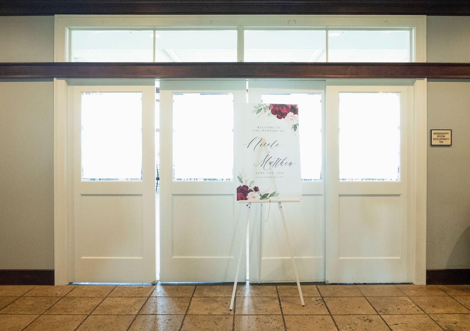 Wedding welcome sign on a white easel displayed at venue entrance doors with floral accents in burgundy and blush.