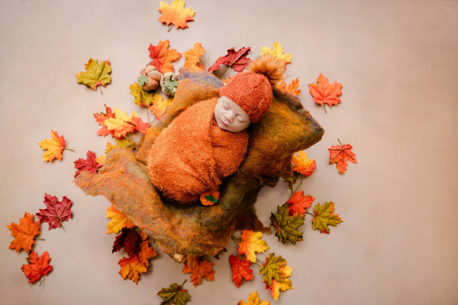 Newborn baby swaddled in orange wrap and hat, sleeping on autumn leaves arrangement for fall newborn photography.