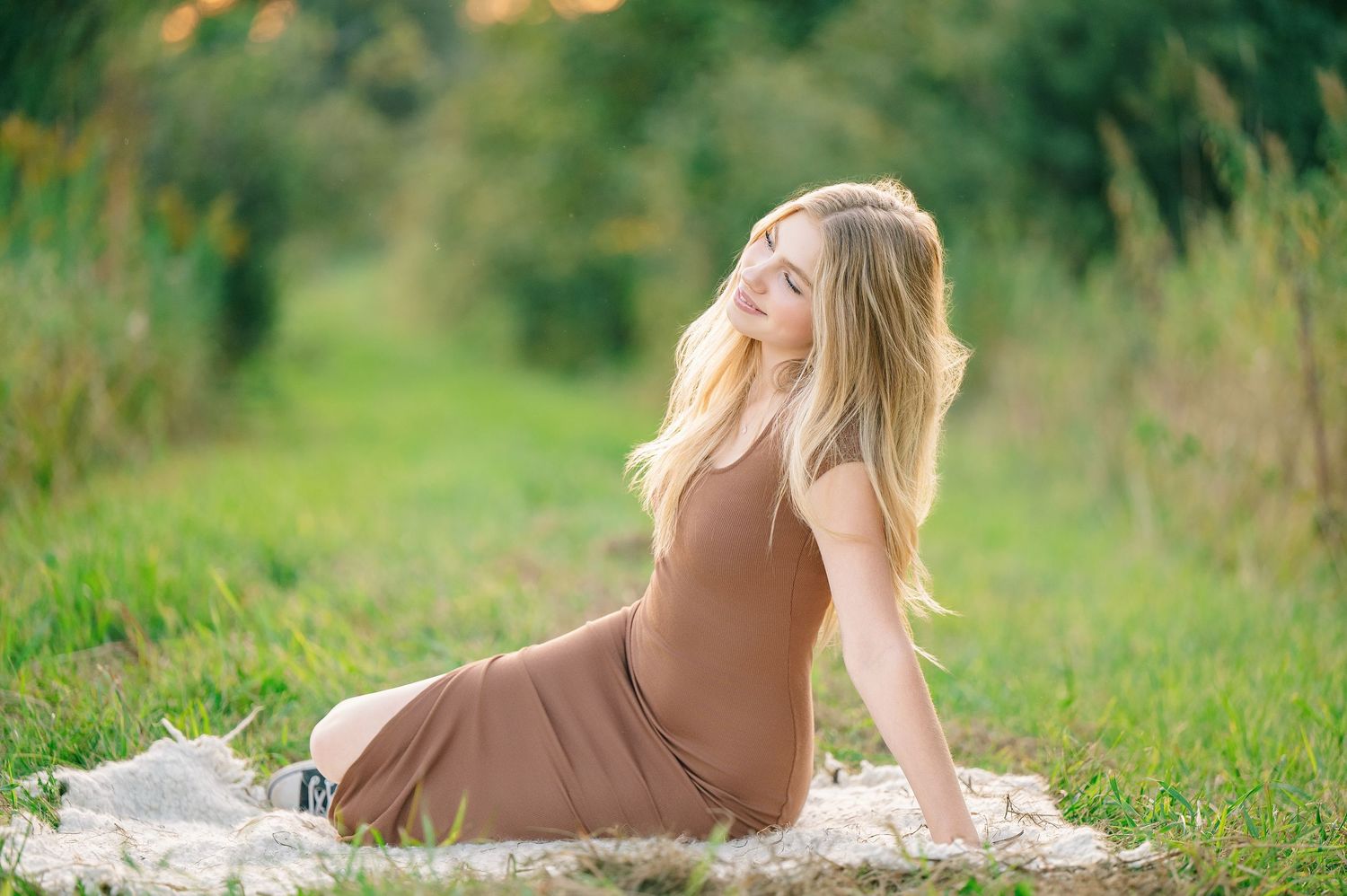 Central MN Senior Photography - Young blonde woman in brown dress sitting gracefully on blanket in lush green meadow during golden hour.