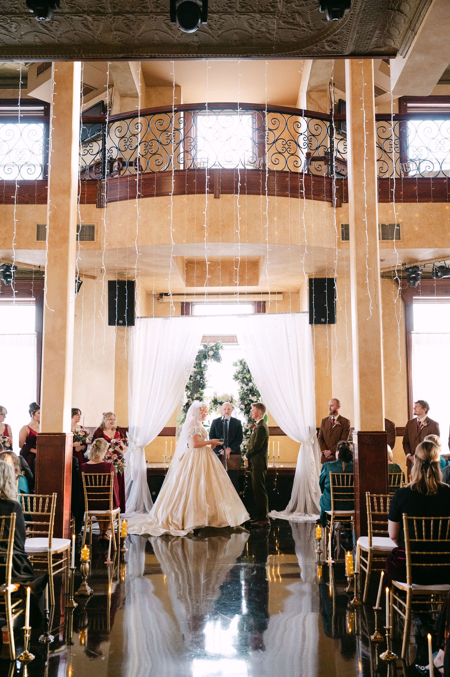 Central MN Wedding Photos | Bride and groom exchange vows at elegant indoor wedding ceremony with draped white curtains and greenery arch.