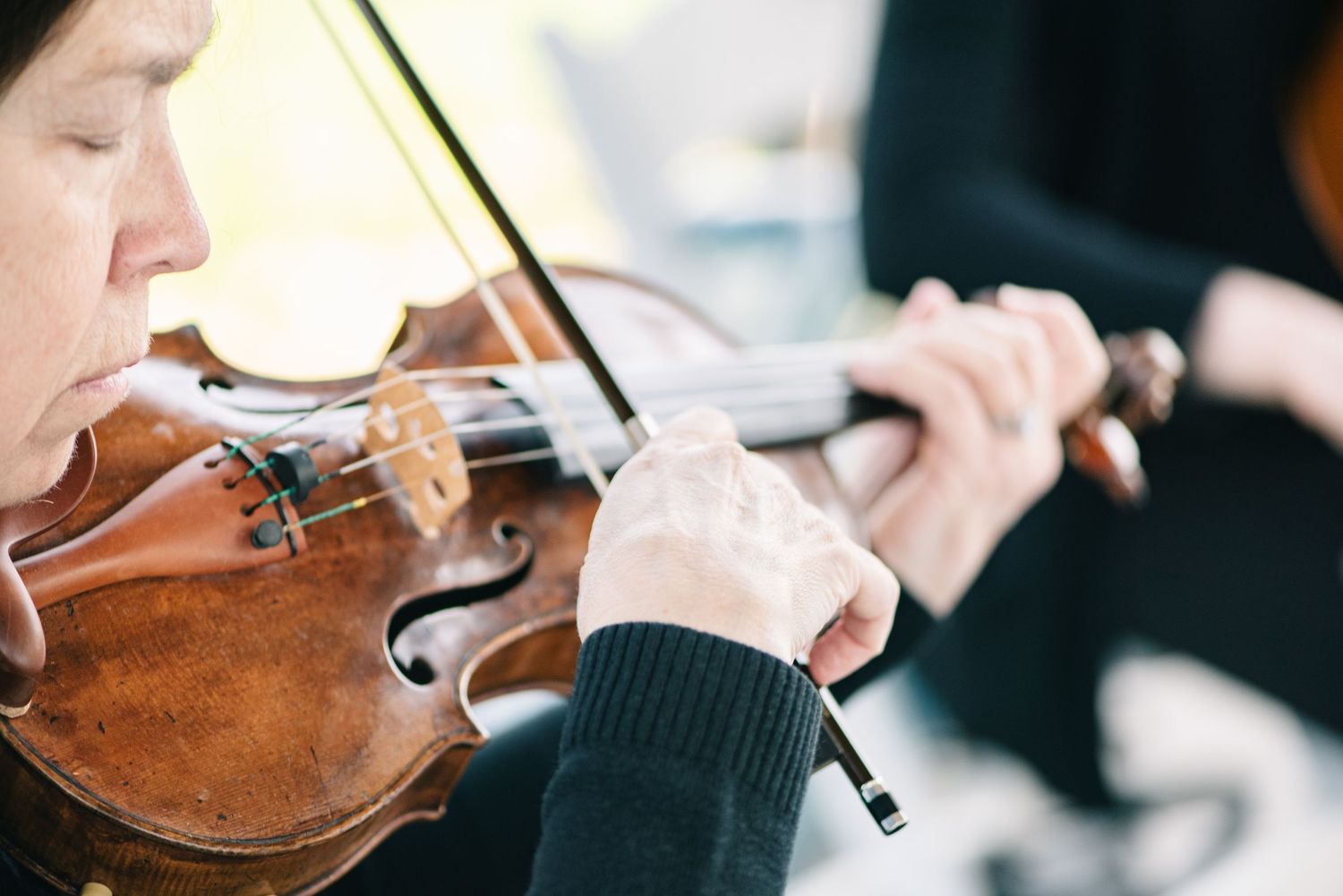 Central MN Wedding Photos -Close-up of a musician playing a wooden violin with a bow, wearing a dark sweater, during an outdoor performance.