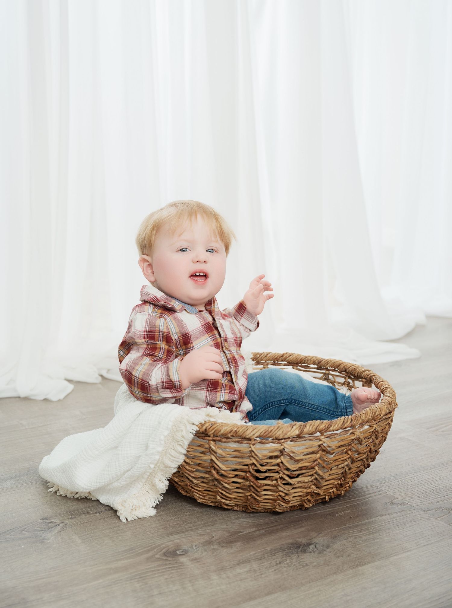 Central MN Milestone Photography - toddler sitting in a wicker basket wearing a plaid shirt and jeans against a white curtain backdrop.