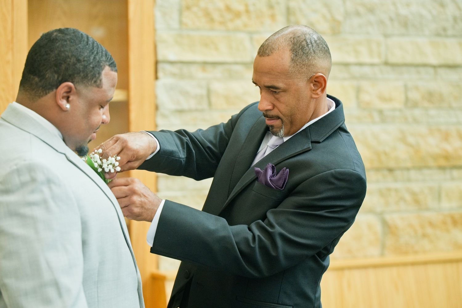 A man in a dark suit pins a boutonniere on a groom in a light gray suit at a wedding ceremony.