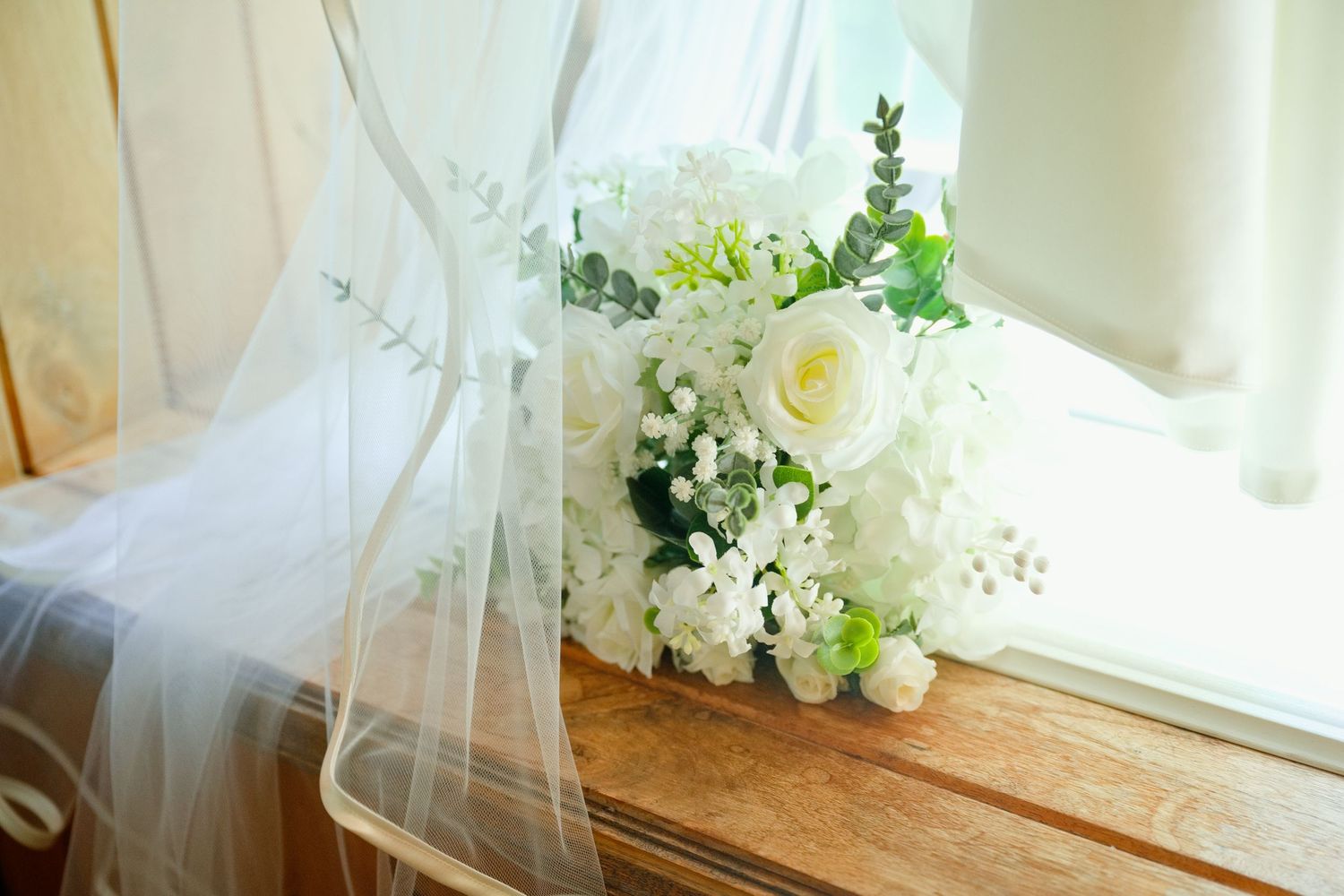 White rose and hydrangea wedding bouquet resting on wooden surface beside sheer bridal veil near sunlit window.