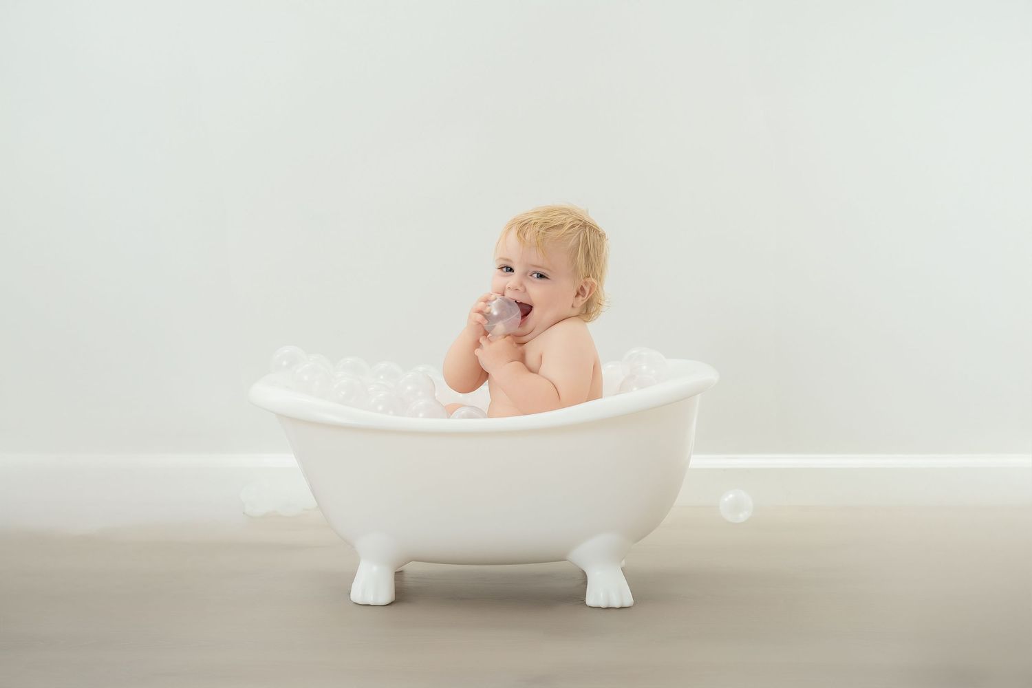 Baby smiles joyfully while sitting in classic white clawfoot bathtub with bubbles during bath time. Princeton MN Milestone, Maternity and Ne