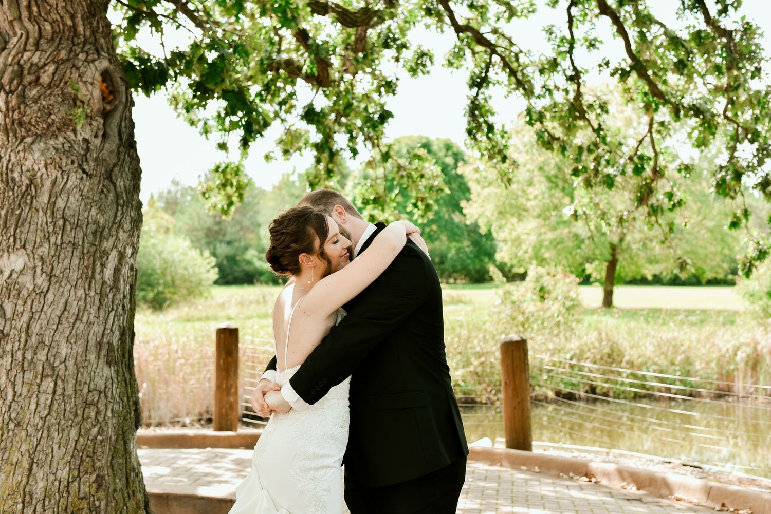 A couple in formal wedding attire dance outdoors under sunlit trees in a romantic garden setting.