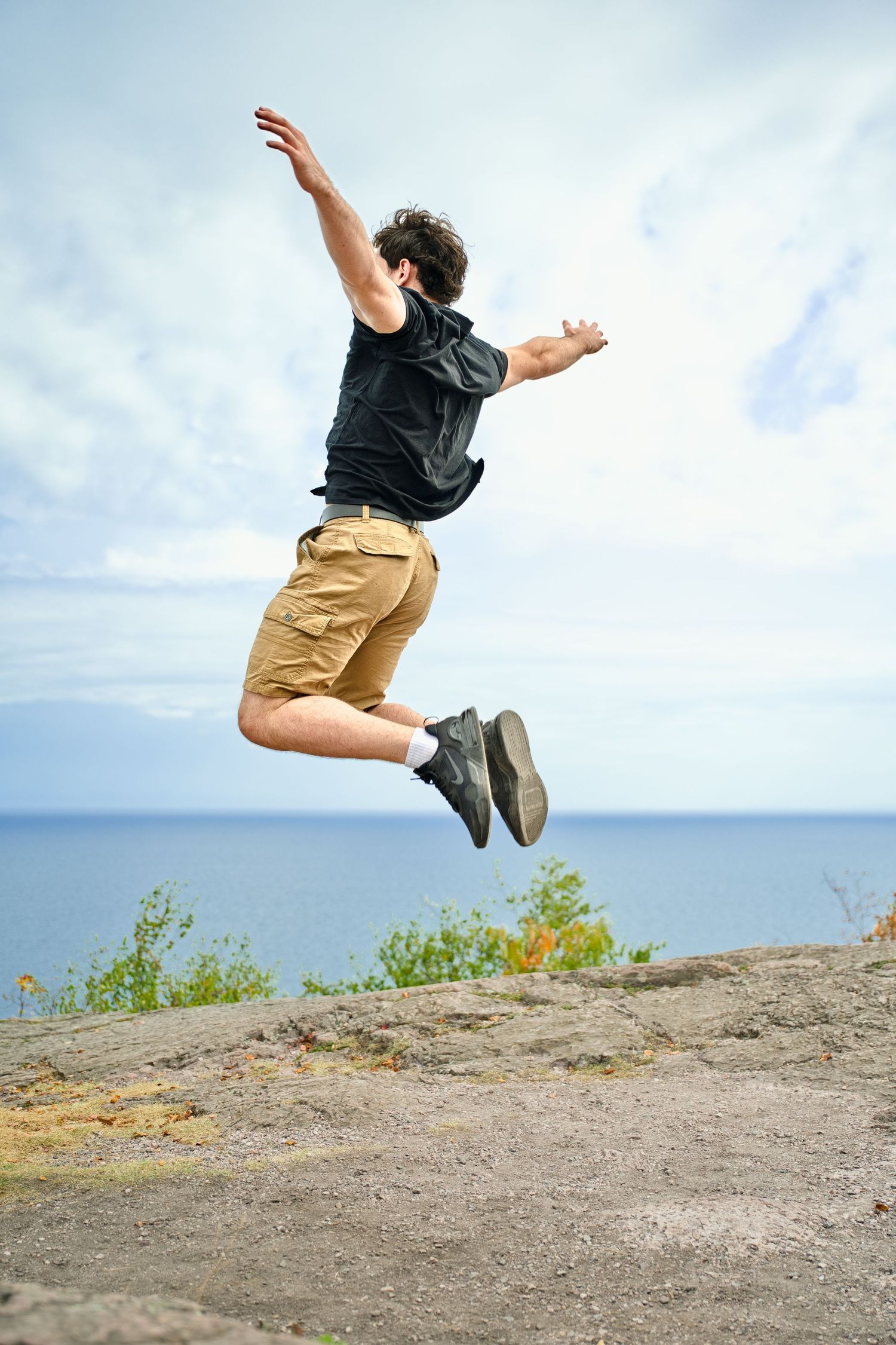 A young man in black shirt and khaki shorts jumping joyfully on rocky cliff with ocean view behind him.