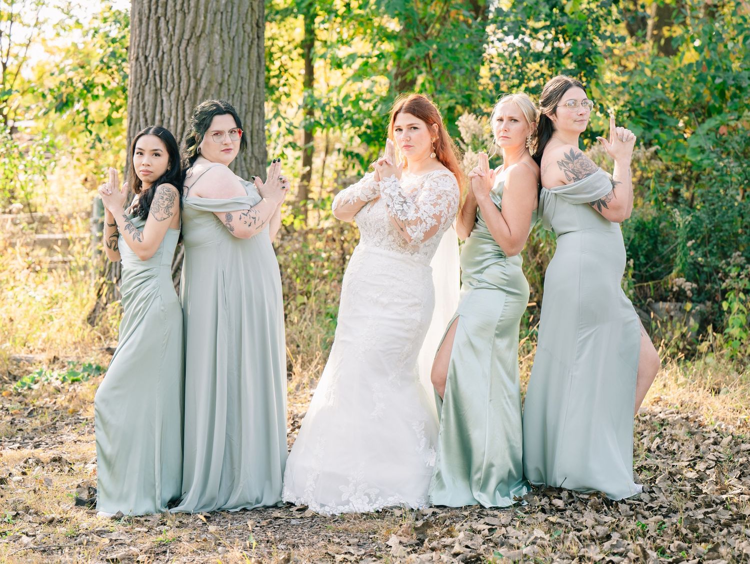 Bride in lace gown poses playfully with four bridesmaids in sage green dresses among autumn trees.