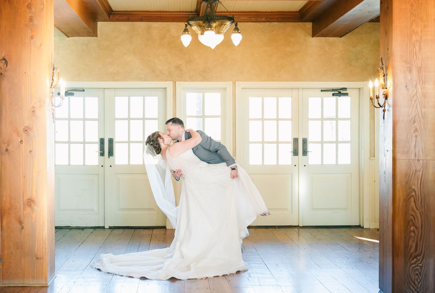 Groom dips bride in elegant wedding venue with white French doors and warm chandelier lighting.