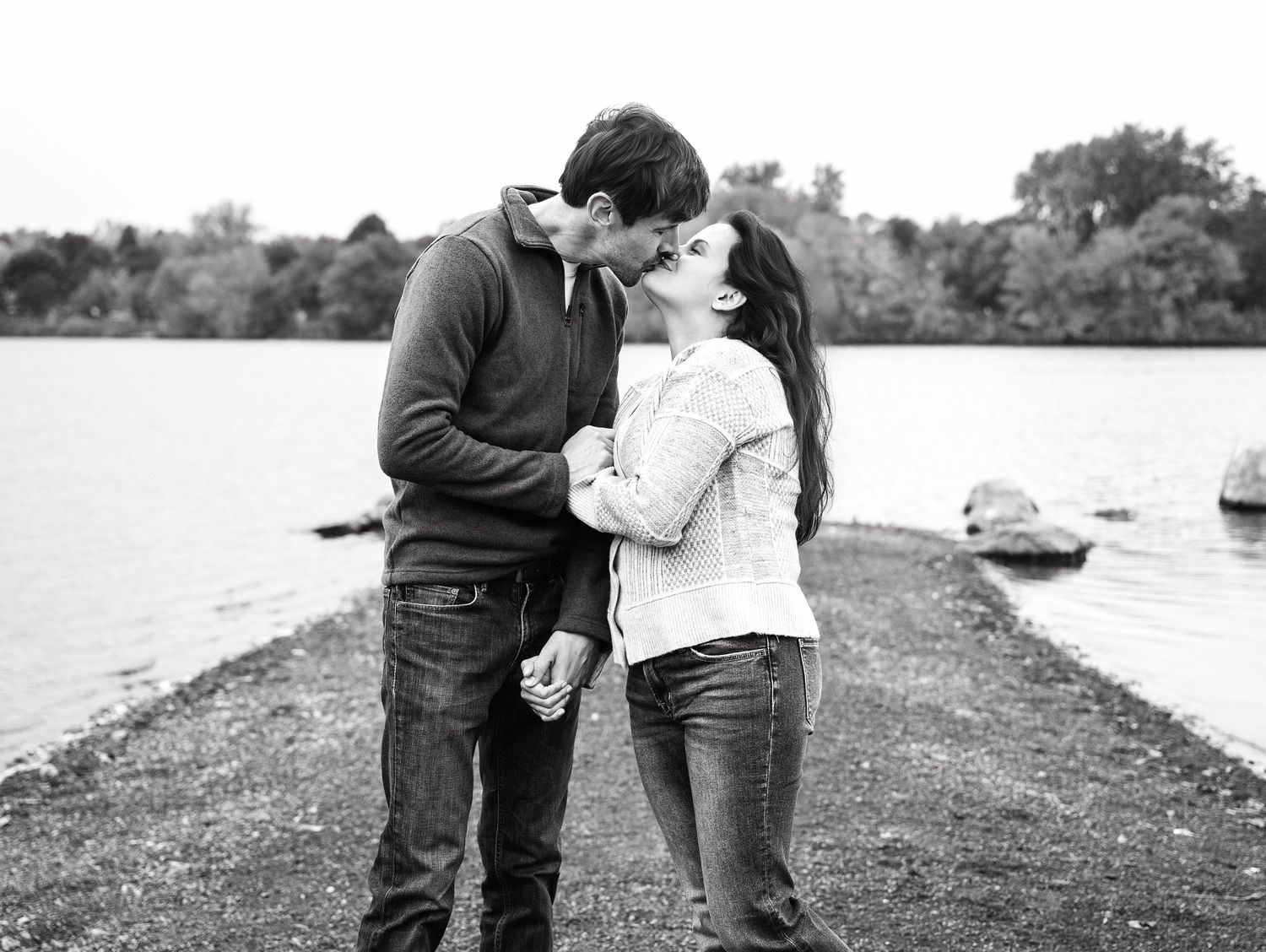 A romantic black and white moment shared between a couple on a lakeside path with boats in the background.