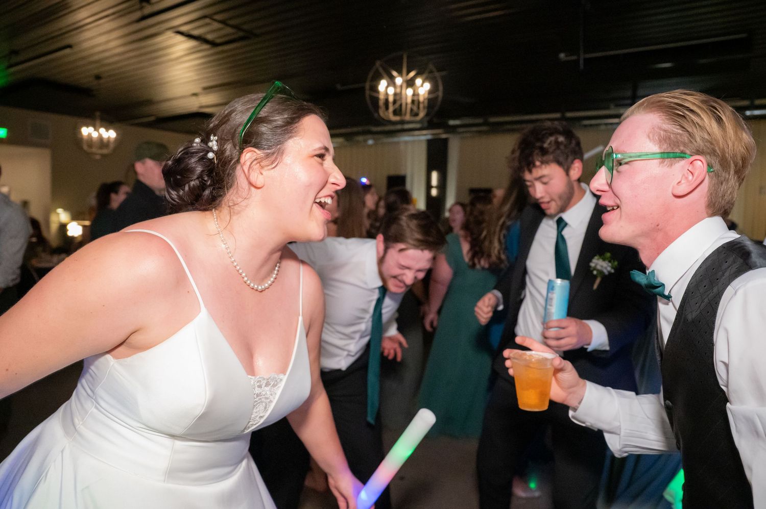 Wedding reception guests dance and celebrate on a dance floor under chandelier lighting at an evening party.