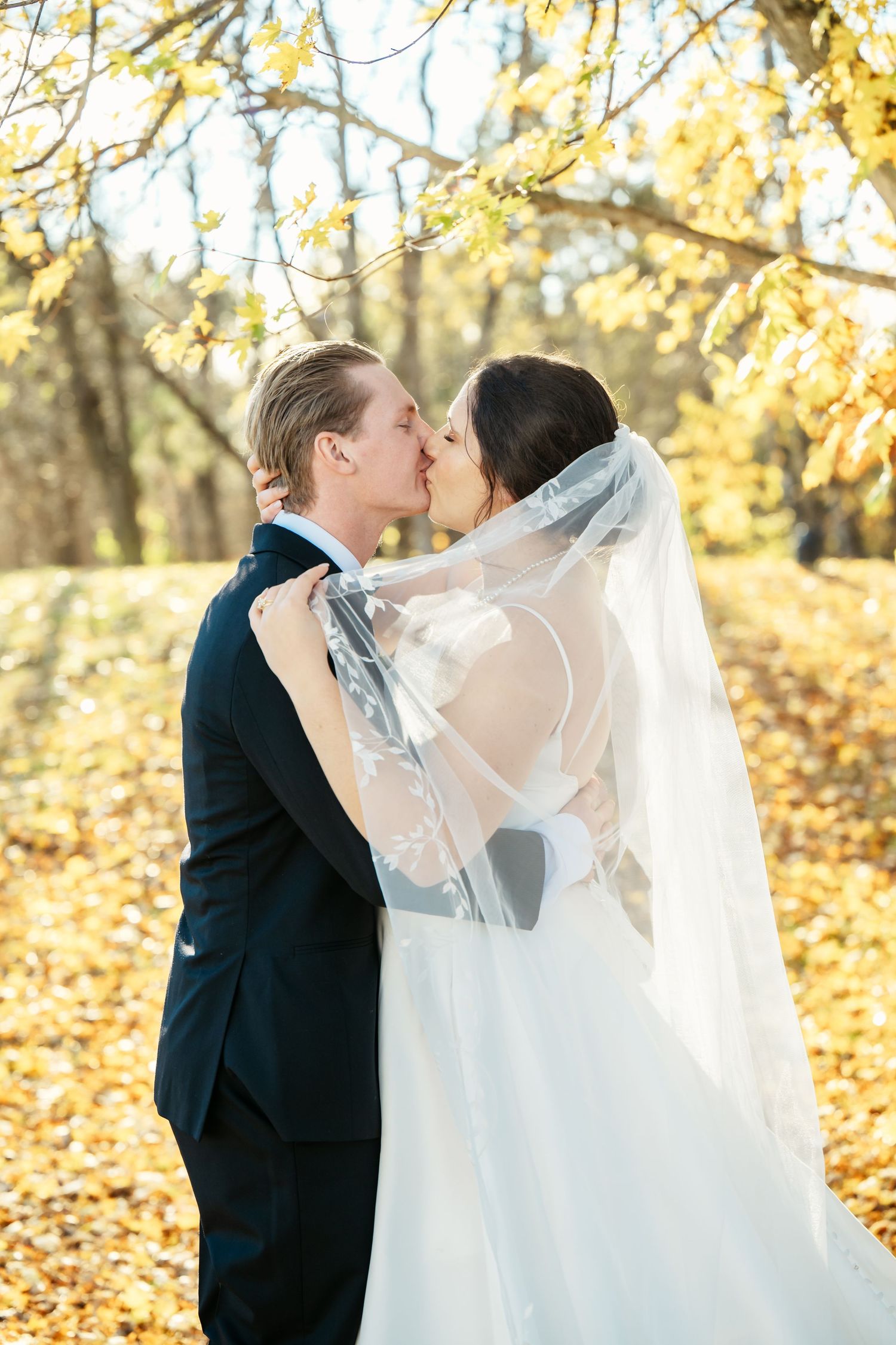 Newlyweds kiss under golden autumn trees with fallen leaves creating a romantic scene.