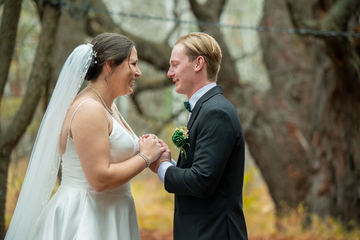 A bride and groom laugh together during their outdoor autumn wedding ceremony surrounded by trees.