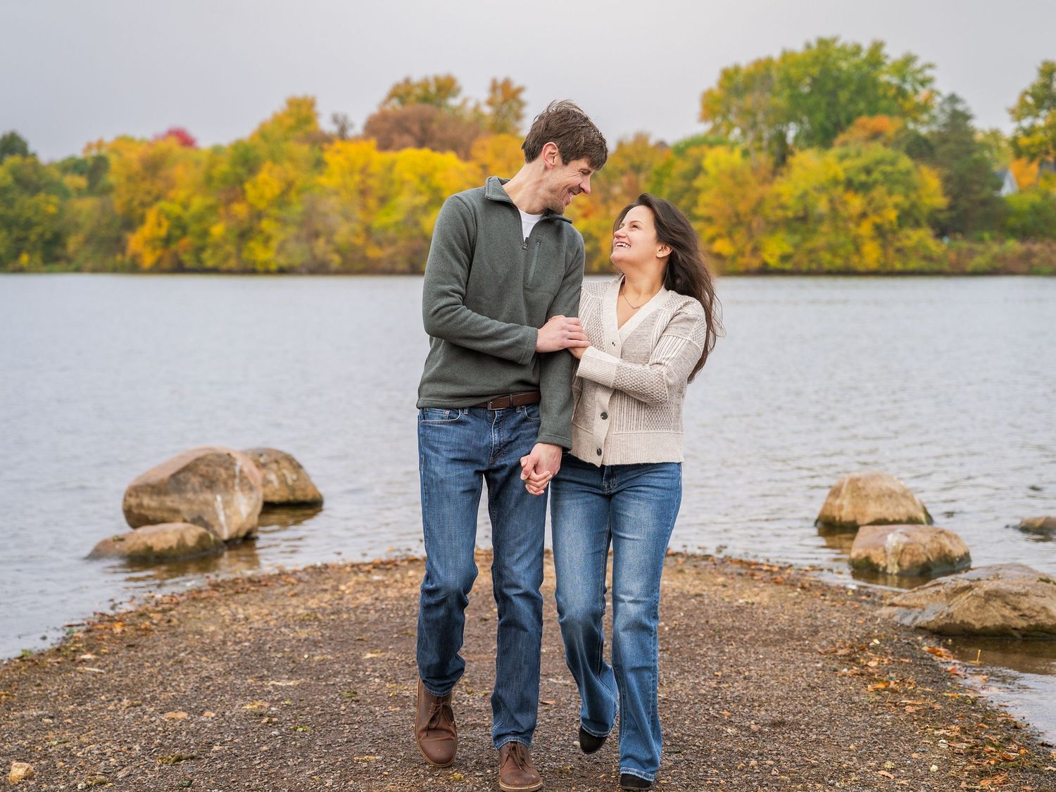 A romantic moment unfolds between a couple standing on a rocky shoreline with autumn trees and a lake in the background at Lake Como MN.