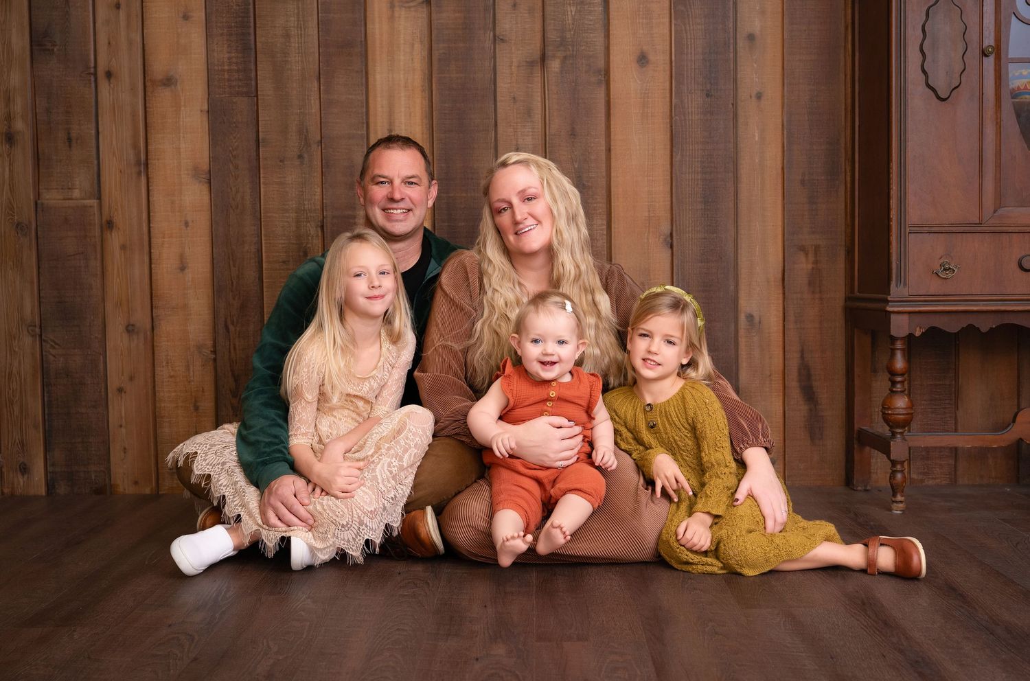 Family poses together in rustic wooden barn setting wearing coordinated fall-colored outfits.