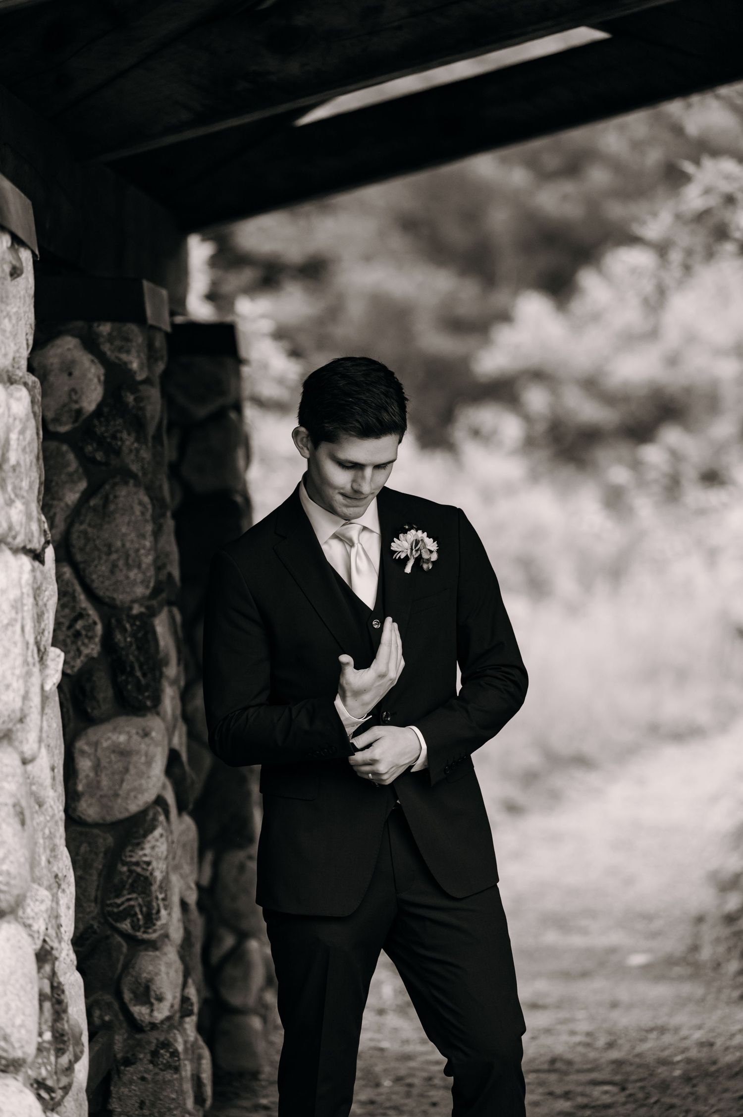 Black and white photo of a groom in formal attire standing against a stone pillar.