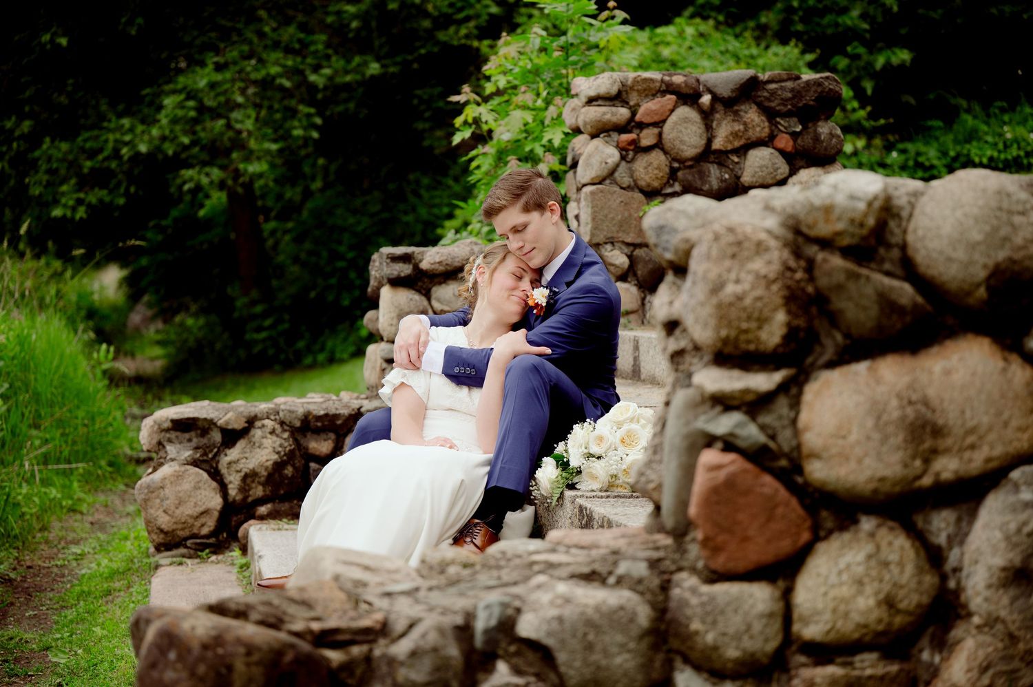 Couple sitting together on rustic stone wall surrounded by lush green forest during outdoor photo session.