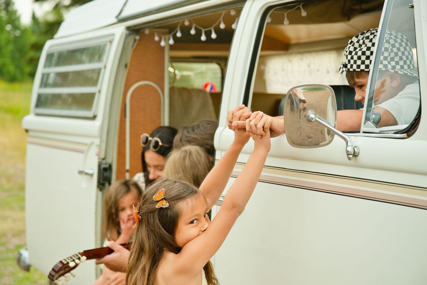 Children playing around a vintage VW camper van, with a kid leaning out the window on a sunny day.
