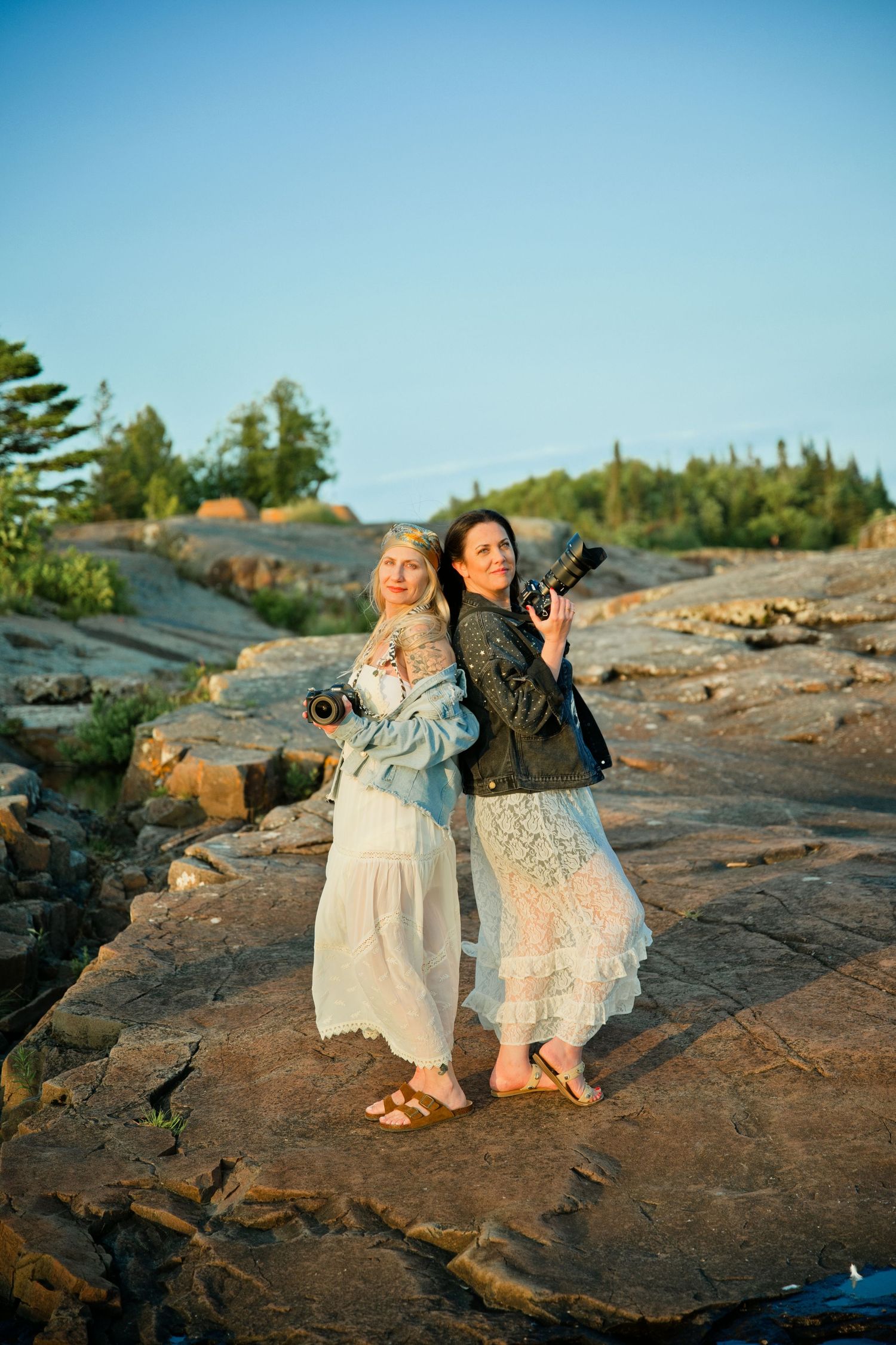 Two women in white lace dresses holding cameras stand back-to-back on rocky shoreline at golden hour.